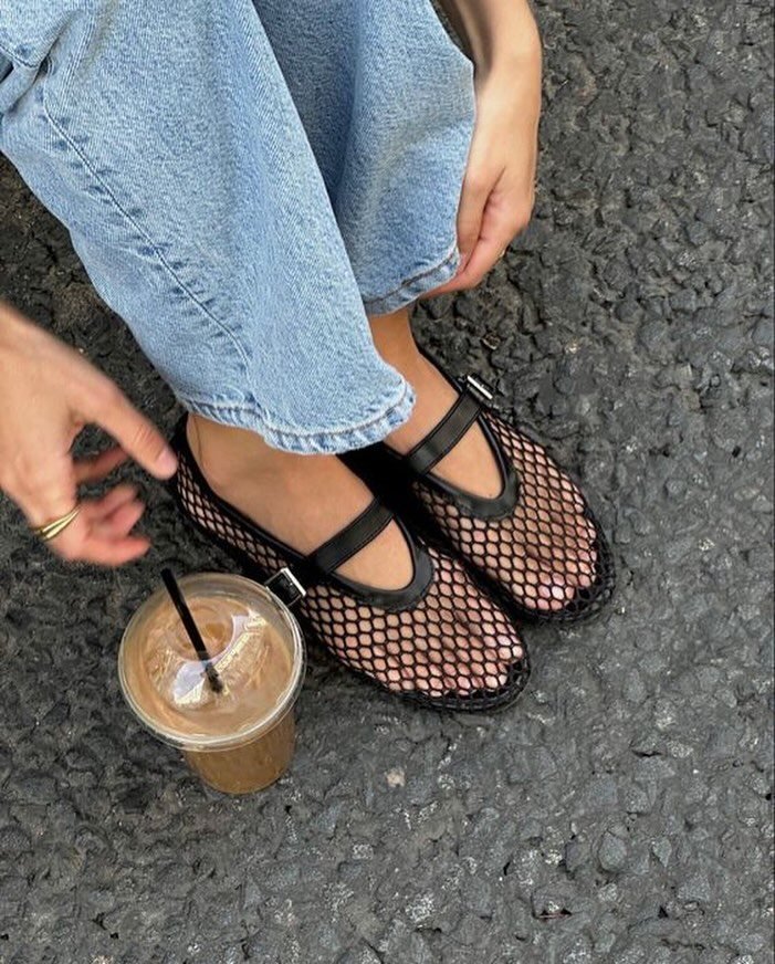 Person wearing black mesh Mary Jane flats and light blue denim jeans sitting near iced coffee.